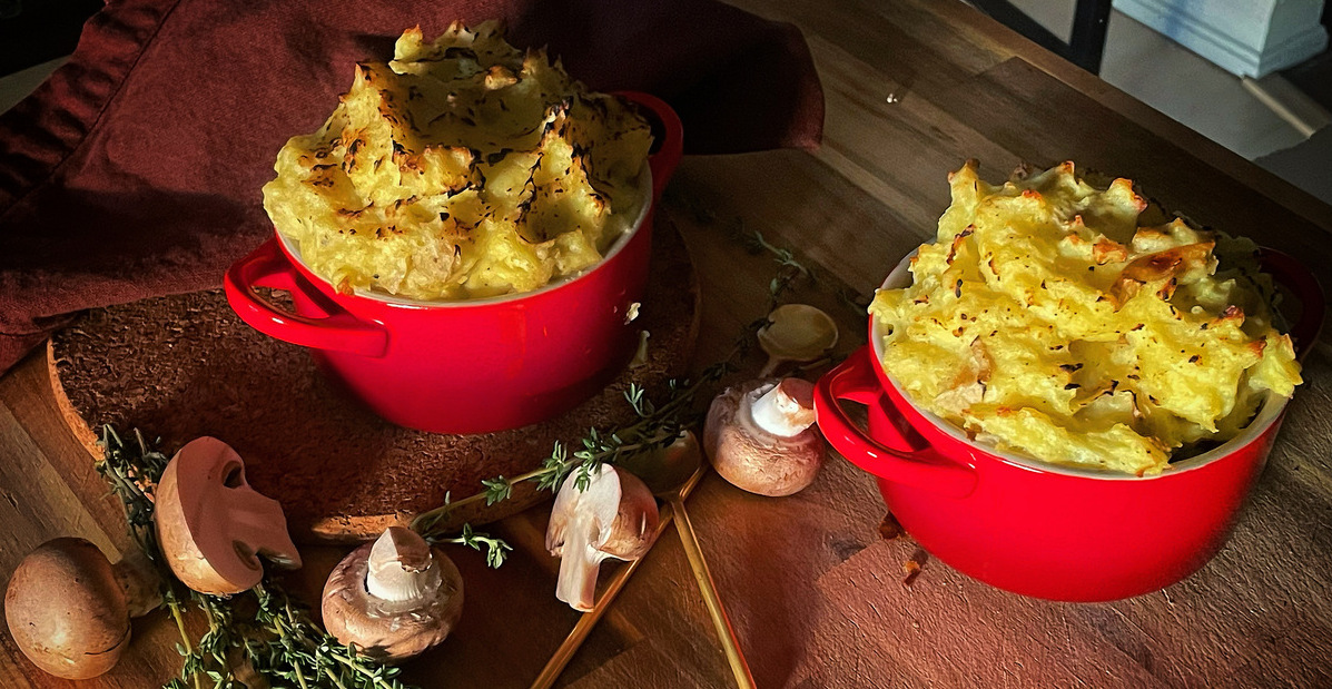 Two red ramekins topped with crusty mashed potato on a wooden board with scattered thyme and mushrooms